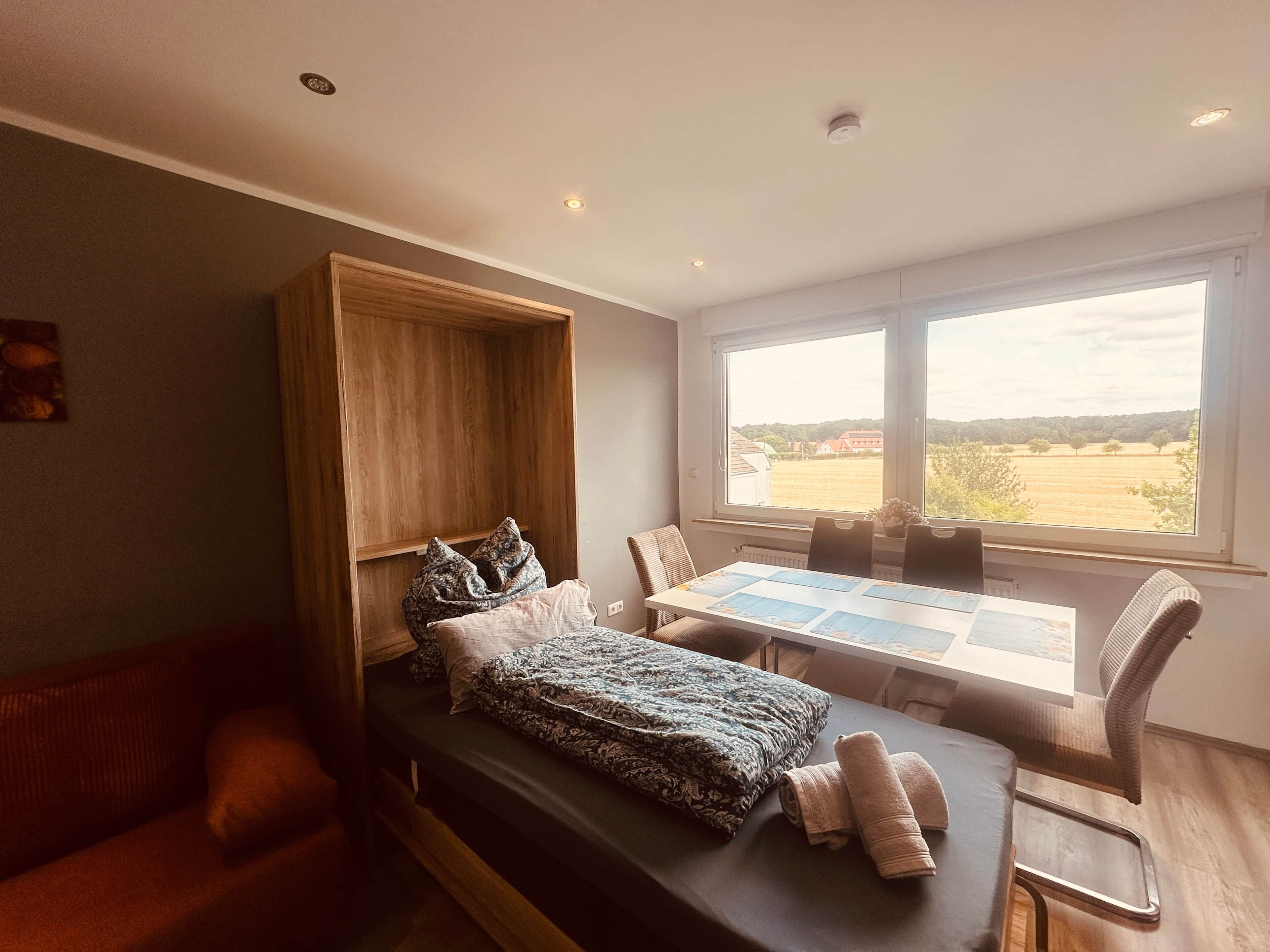 Une chambre à coucher moderne, avec un lit, une armoire en bois, une table à manger avec des chaises et une grande fenêtre donnant sur la campagne.