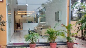 A modern living room with a glass door leading to a kitchen and dining area. There are potted plants outside and a visible water pipe on the left. - Hotel PSR Executive Rooms by VINHOTELS (Bengaluru)