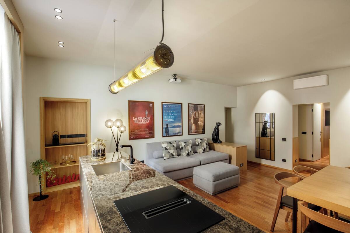 Una sala de estar moderna con sofá, reposapiés y comedor. Hay carteles en la pared y una lámpara de techo.