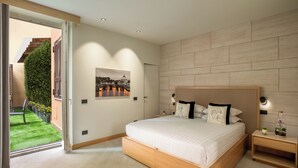 A modern bedroom with a large bed, a wooden headboard, a nightstand, and a view of a green garden through a sliding door. - Ripetta Luxury Apartments (Rome)