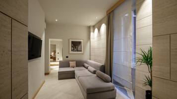 A modern living room with a sectional sofa, a flat-screen TV mounted on the wall, and a large window with blinds.