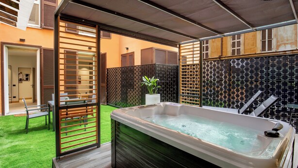 A modern outdoor hot tub area with a wooden deck, a small seating area, and a patterned wall. - Ripetta Luxury Apartments (Rome)