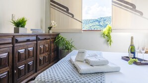 A hotel room with a bed, a wooden dresser, a window with a view of a green landscape, a potted plant, and a tray with a bottle and glasses. - Apartamento aconchegante. Vista incrível (Águas de Lindóia)