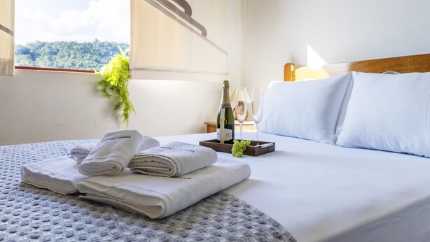 A neatly made bed with white linens, a tray with a bottle and glasses, and a window with a view of greenery. - Apartamento aconchegante. Vista incrível (Águas de Lindóia)