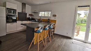 A modern kitchen with a central island, bar stools, and a sliding glass door leading to a patio. - Maison Familiale Entre mer et Centre-ville (Ondres)