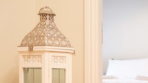Un mueble blanco con puertas de vidrio y una parte superior decorativa, situado en una habitación con suelo de madera y una cama al fondo.
