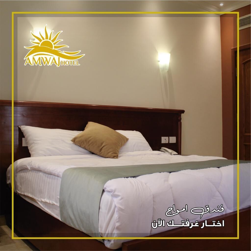Habitación de hotel con una cama de madera, ropa blanca, una almohada color beige y una lámpara fijada a la pared.