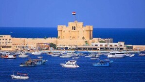 A coastal town with a prominent castle-like structure, a marina with various boats, and buildings along the waterfront. - Amwaj Hotel (Alexandria)