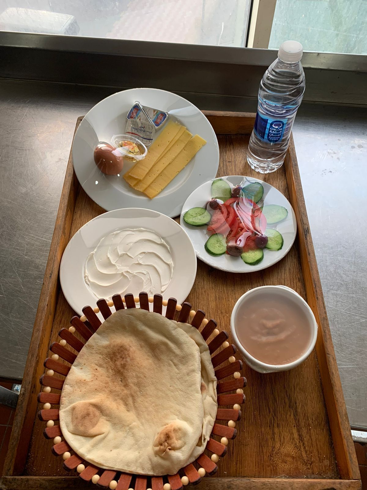 Een dienblad met een bord met plakjes komkommer, tomaten en rode uien, een bord met plakjes brood, een schaal hummus, een fles water, en een bakje ongezouten boter.