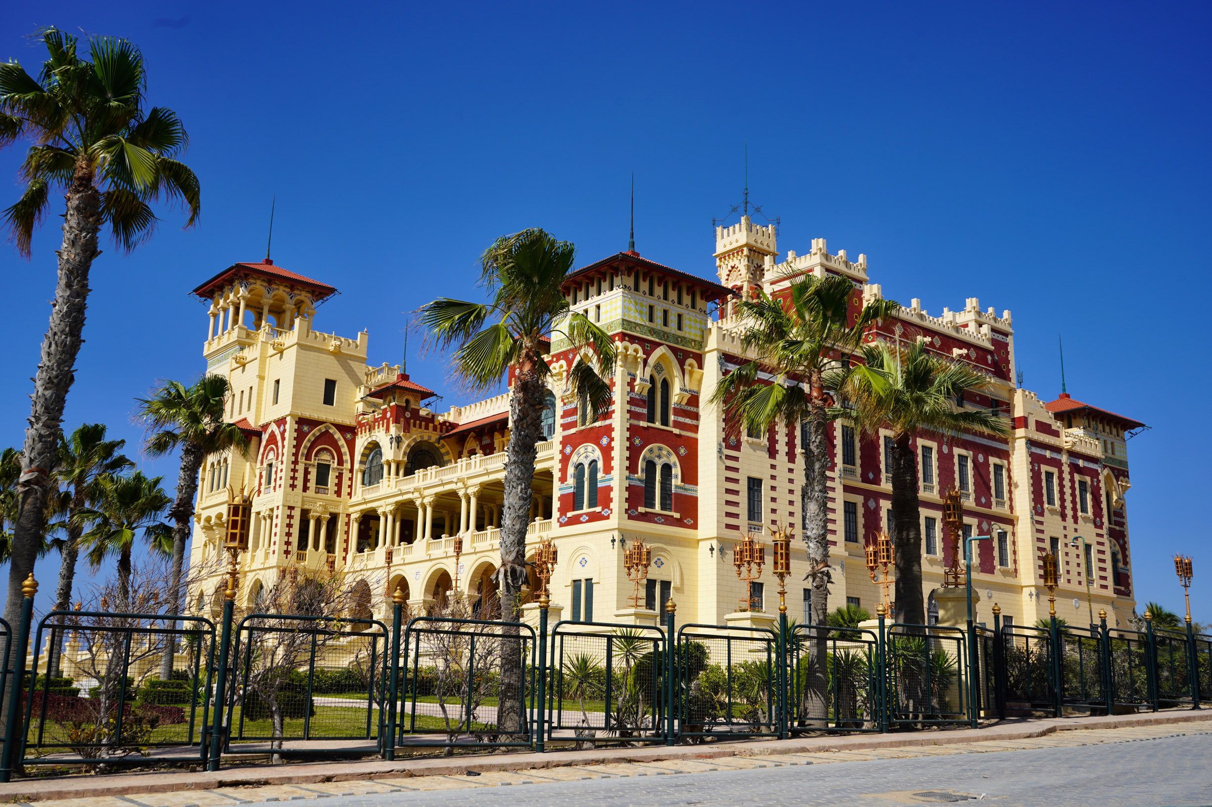 Un edificio grande y ornamentado con múltiples torres y balcones, rodeado de palmeras y una valla.