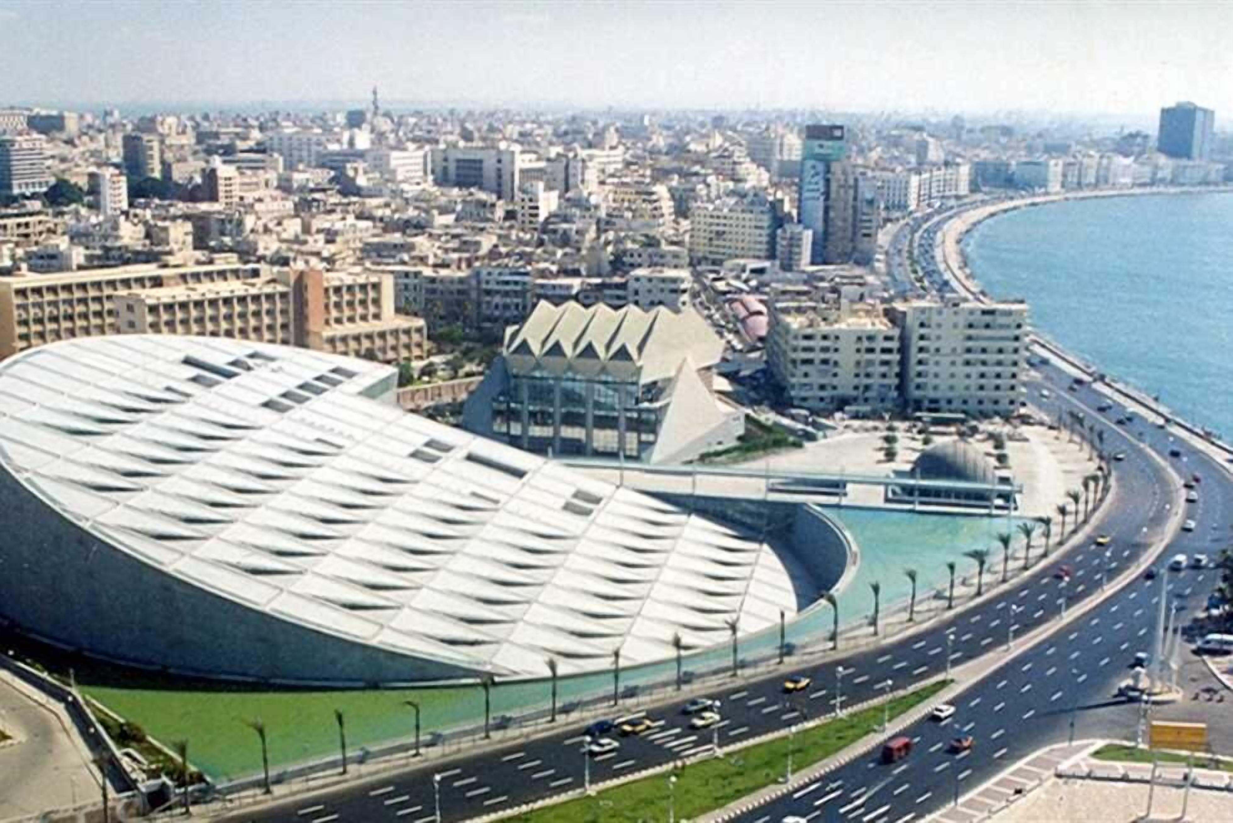 A coastal cityscape with a prominent curved building near the waterfront, a busy road, and surrounding urban buildings.