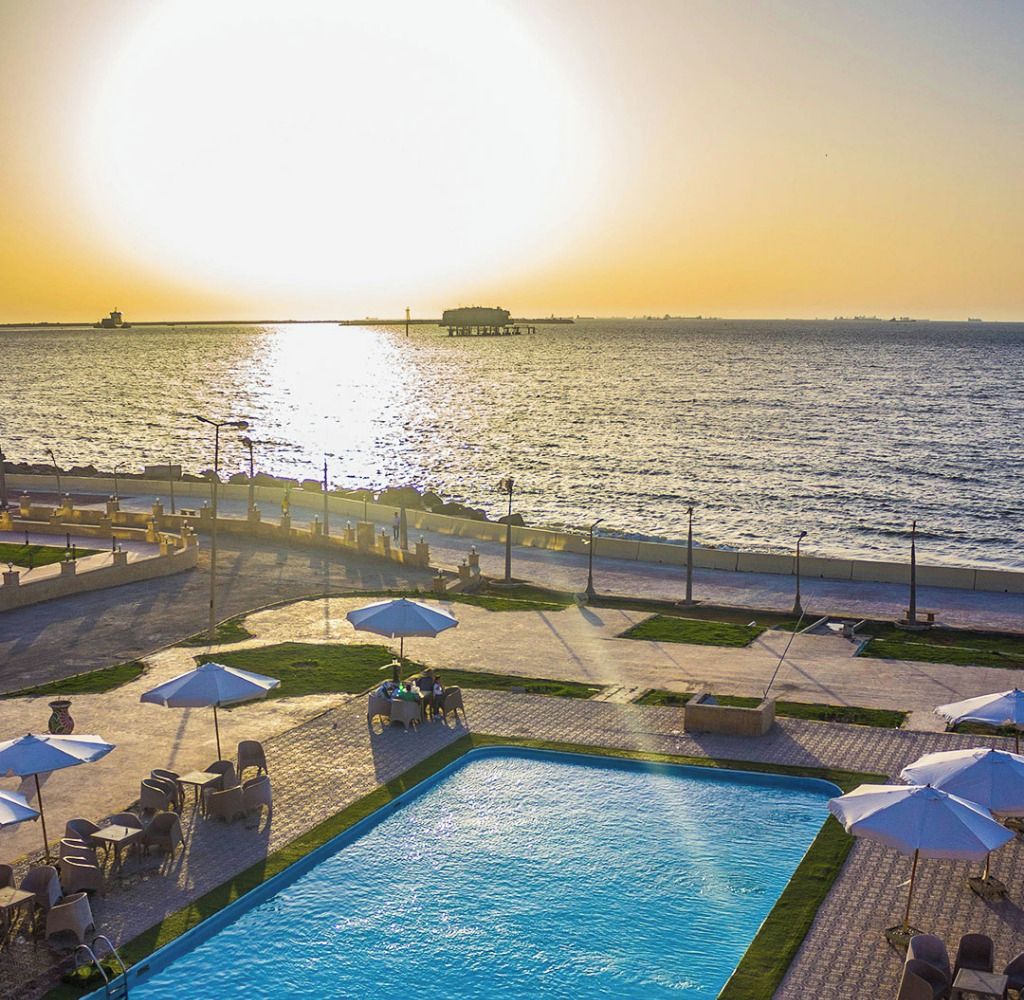 A poolside area with a view of the sea at sunset, outdoor seating, and a sign reading "START YOUR Summer Camp".