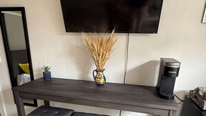 Ett modernt vardagsrum med ett mörkt träbord, två vadderade bänkar, en vas med torkade växter, en kaffebryggare och en platt-TV som är monterad på väggen.
