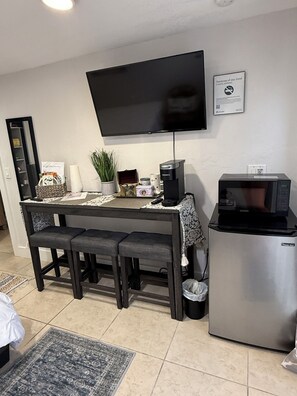 A compact living space with a kitchenette, a flat-screen TV mounted on the wall, a small refrigerator, and a dining area with a table and bench.