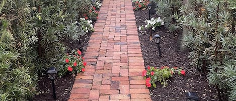 Un sendero de ladrillo que lleva a una puerta blanca con un diseño de celosía, rodeada de follaje y flores rojas.