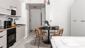 A modern kitchen with a microwave, oven, and sink. There is a dining area with a table set for four, surrounded by chairs. A bed is visible in the background. - Prime Apartments Pori Central (Pori)