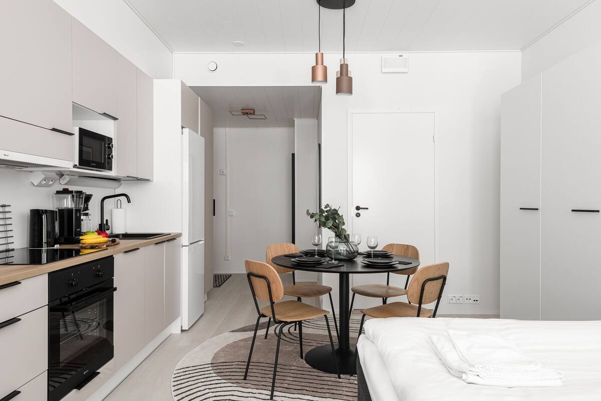 Eine moderne Küche mit Mikrowelle, Backofen und Spüle. Ein Essbereich mit Tisch für vier Personen, umgeben von Stühlen. Im Hintergrund ist ein Bett zu sehen.