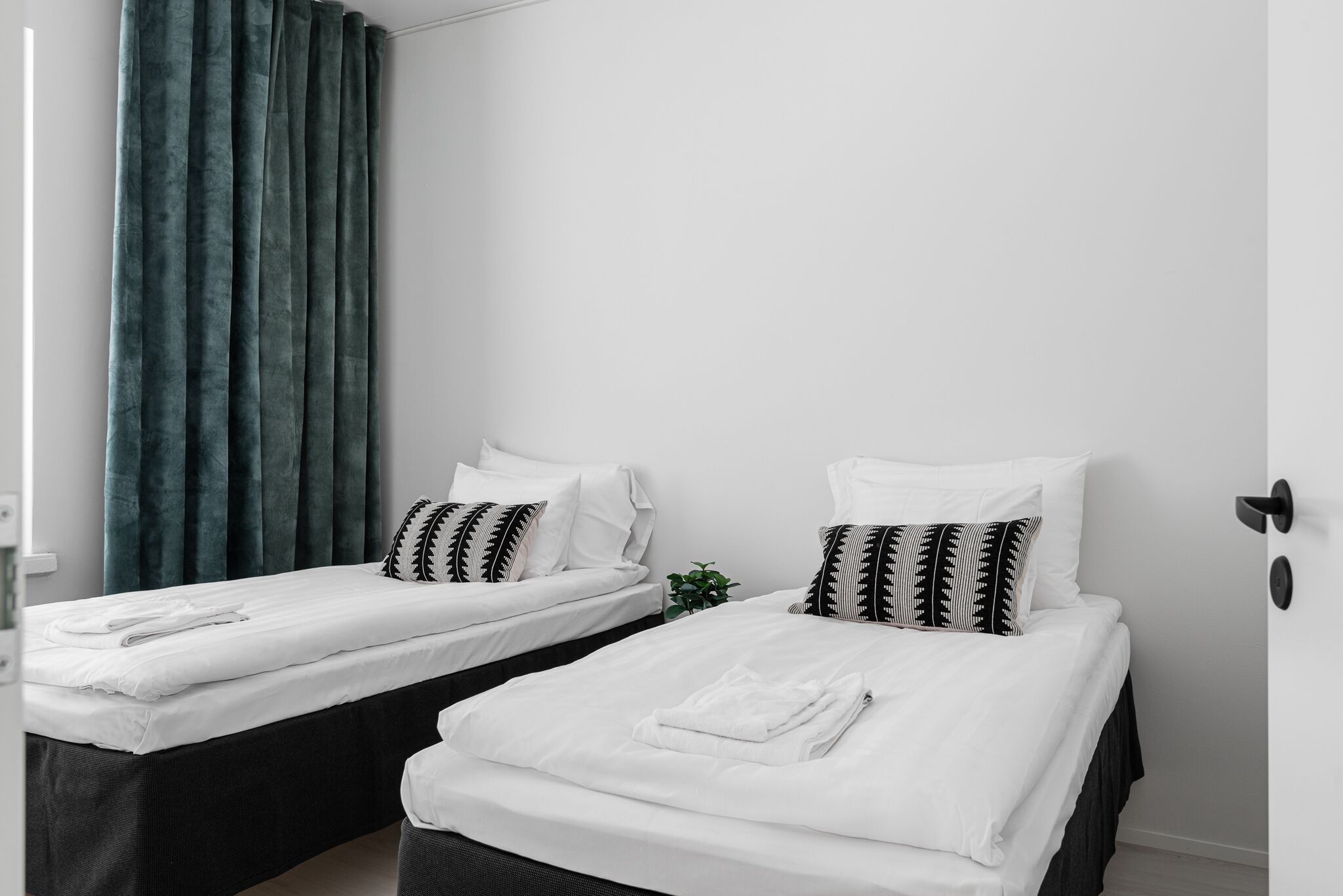 Two single beds with white linens and black and white patterned pillows, a dark green curtain, and a small potted plant.