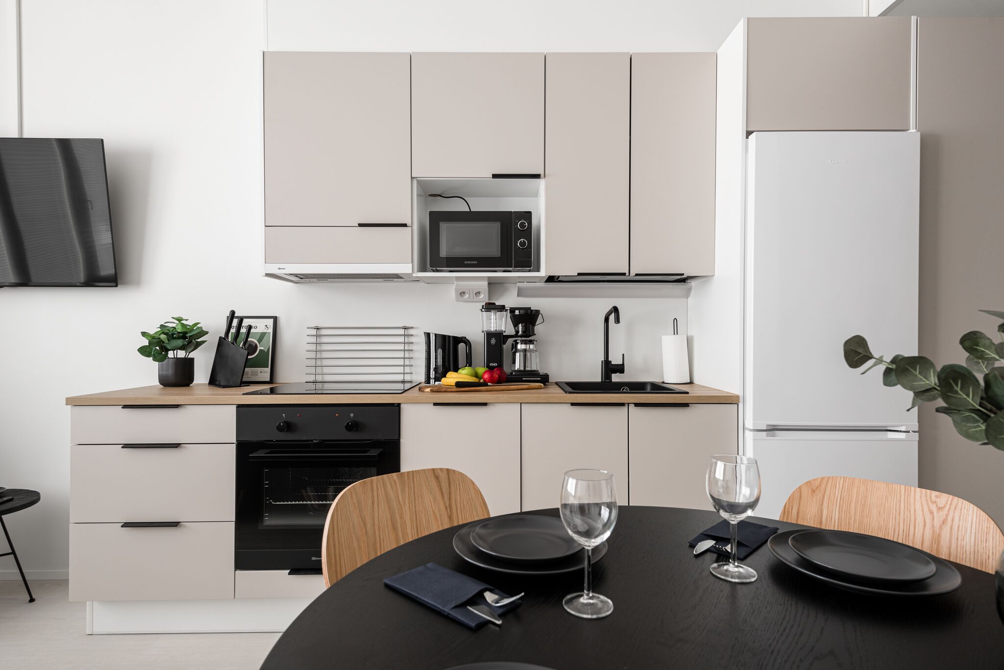 A modern kitchen with a microwave, oven, and refrigerator. There is a dining table with chairs and a wine glass.