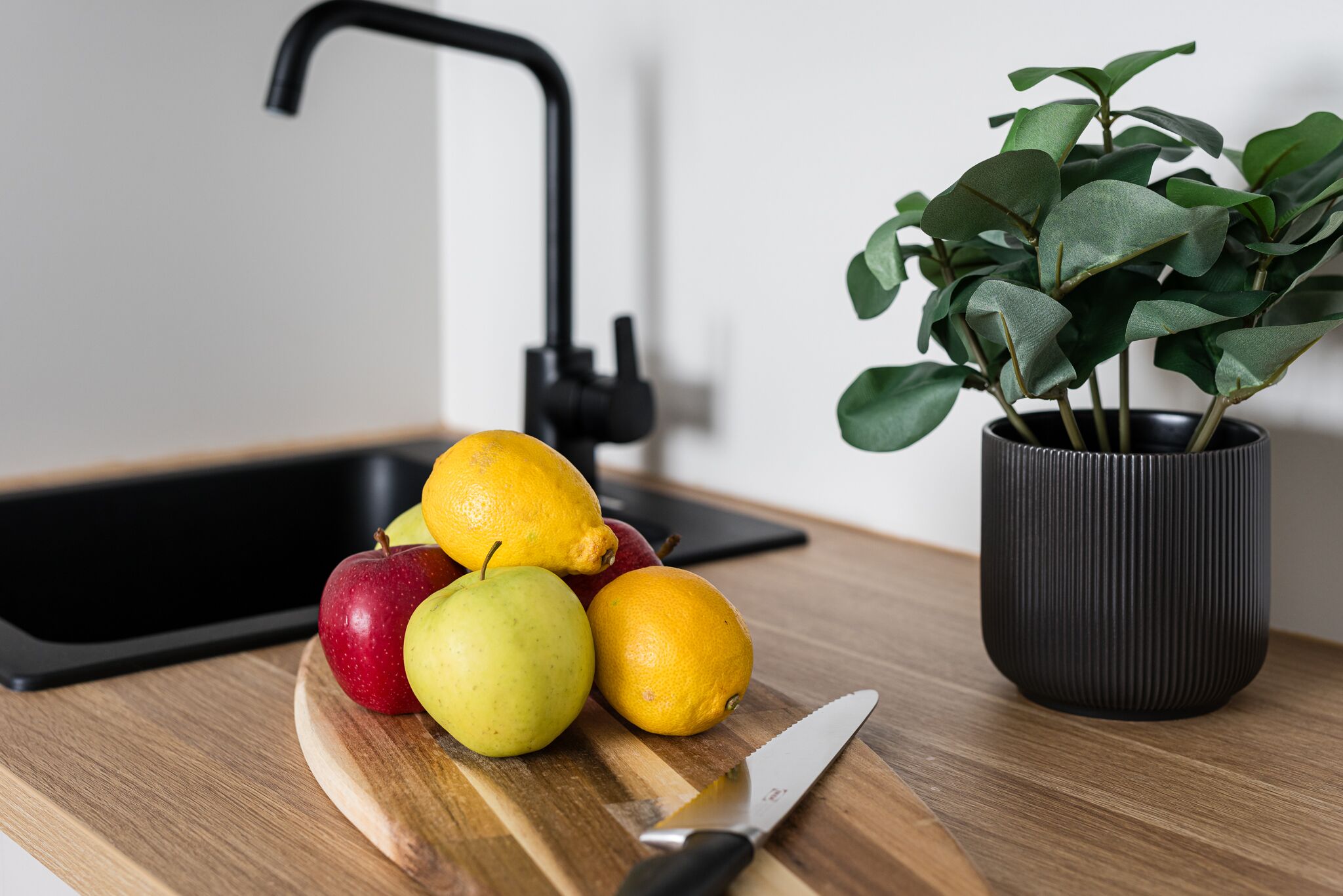A kitchen counter with a cutting board, fruits, a knife, a potted plant, and a black faucet.