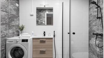 A modern bathroom with a washing machine, wooden cabinet, mirror, and shower.