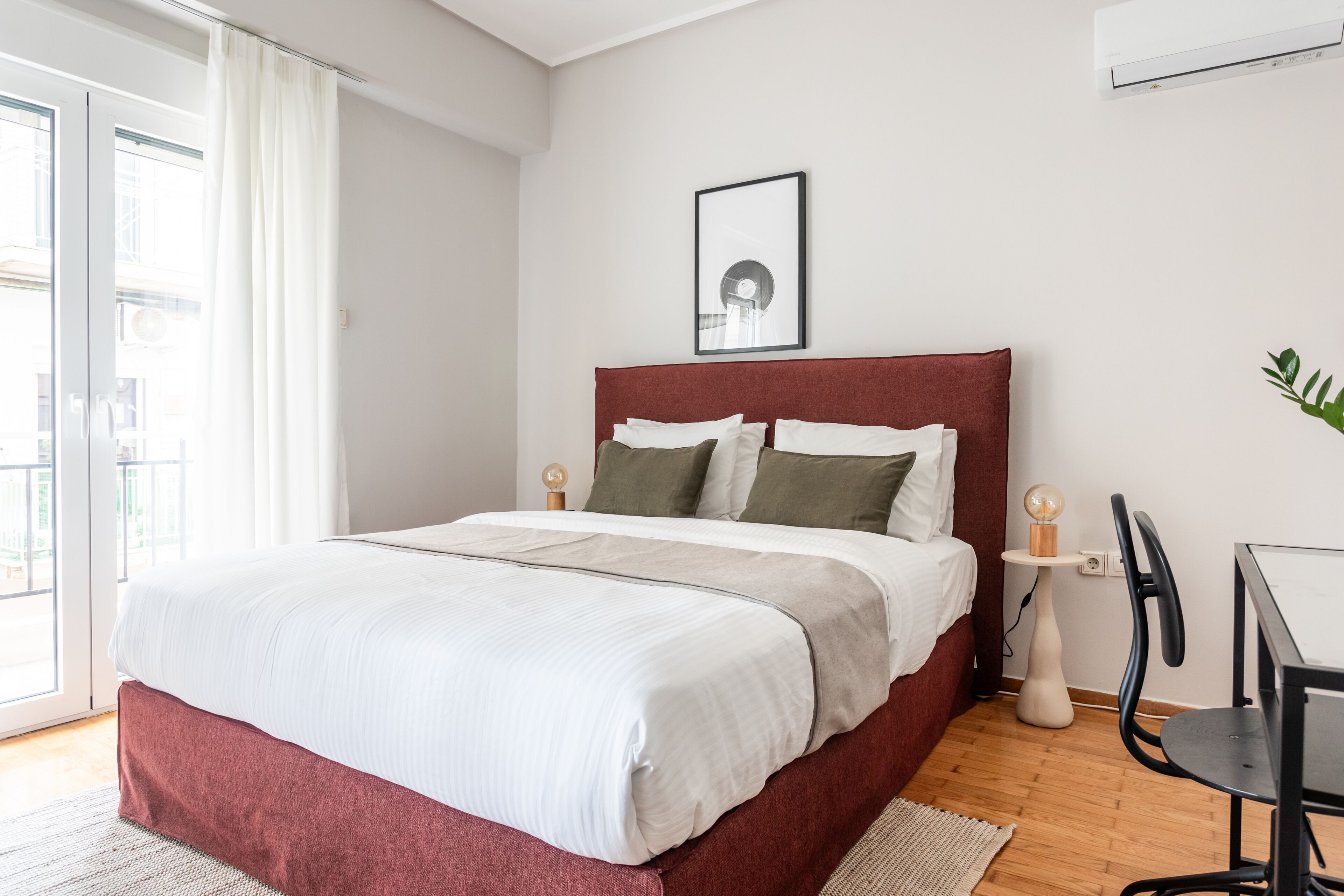 Une chambre à coucher avec un lit, une table de chevet, une chaise, un tableau encadré au mur et une fenêtre avec des rideaux.