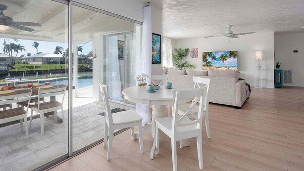 A modern living room with a glass wall, a round dining table with white chairs, a beige sofa, and a ceiling fan.