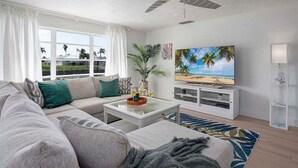 A modern living room with a large window, a ceiling fan, a flat-screen TV, and a cozy seating area.