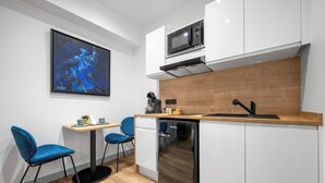 A compact kitchen with a microwave, sink, and dishwasher. There is a small table with chairs and a painting on the wall. - L'App-Art (Nice)