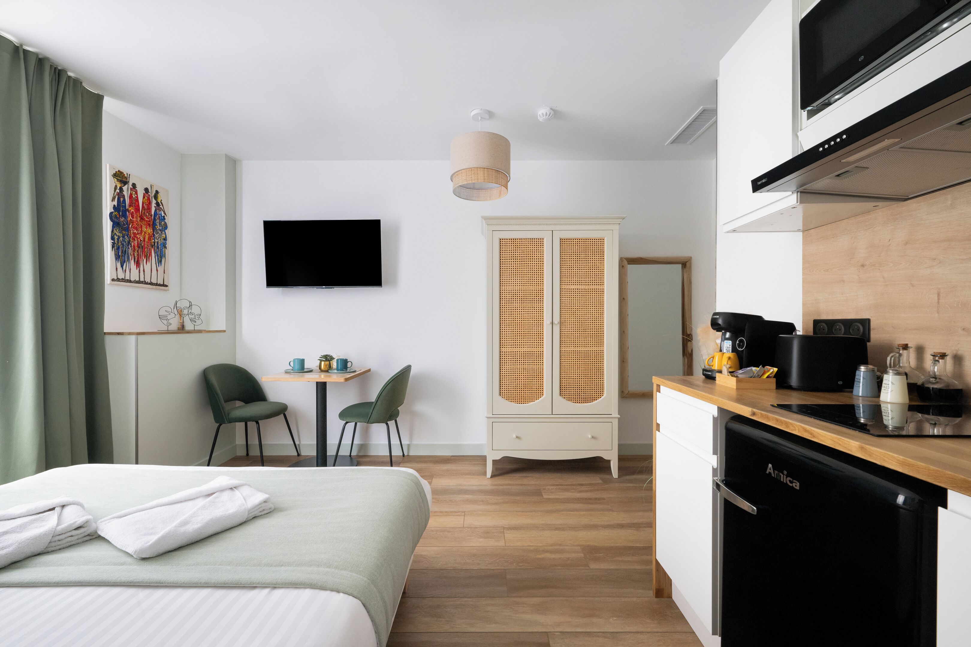 Ett hotellrum med en säng, ett skrivbord med stolar, en tv, en garderob och ett litet köksområde med diskmaskin.
