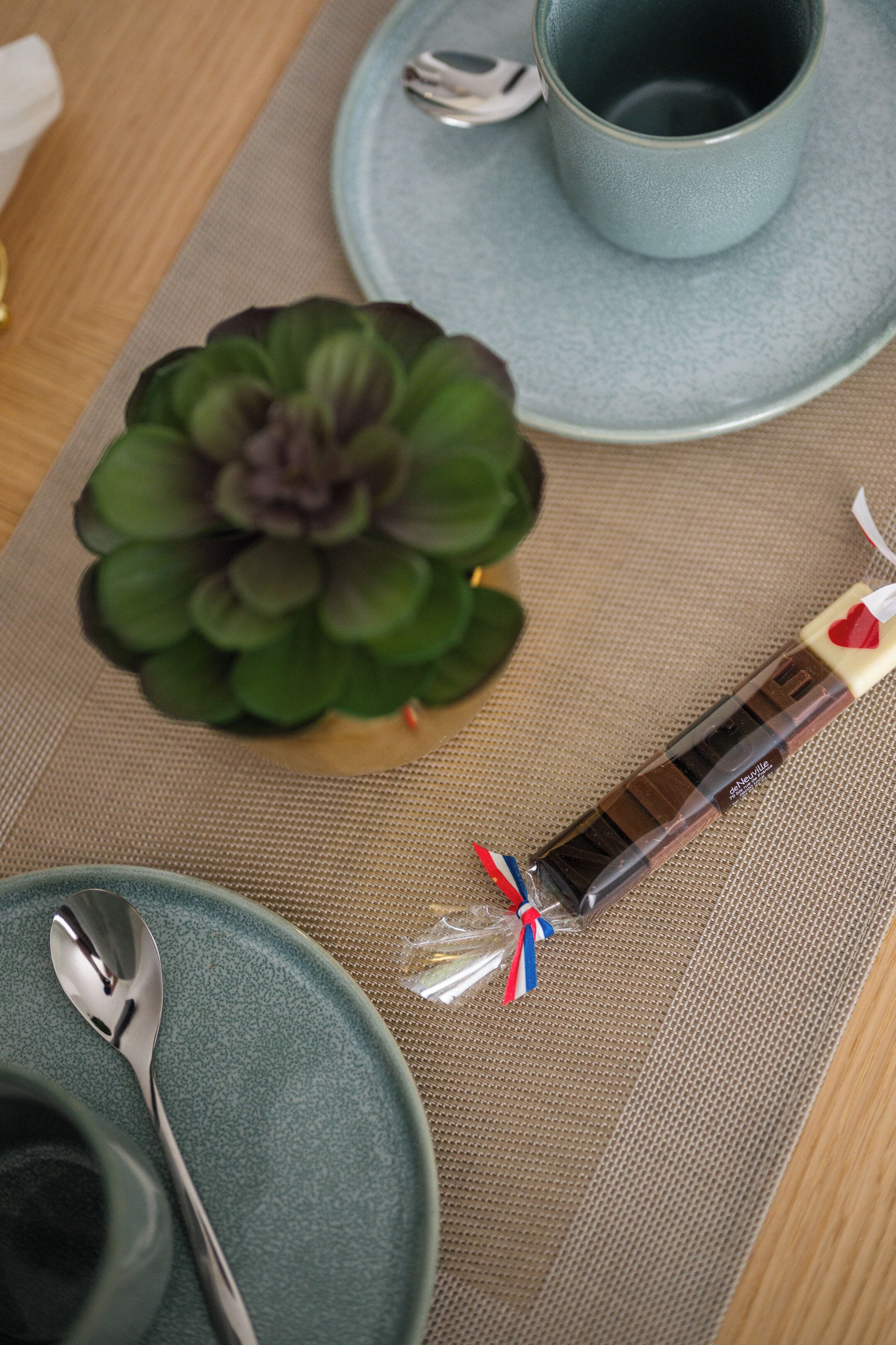 Une table avec un set de table, une petite plante succulente verte, une barre de chocolat emballée, une tasse bleue et une cuillère.