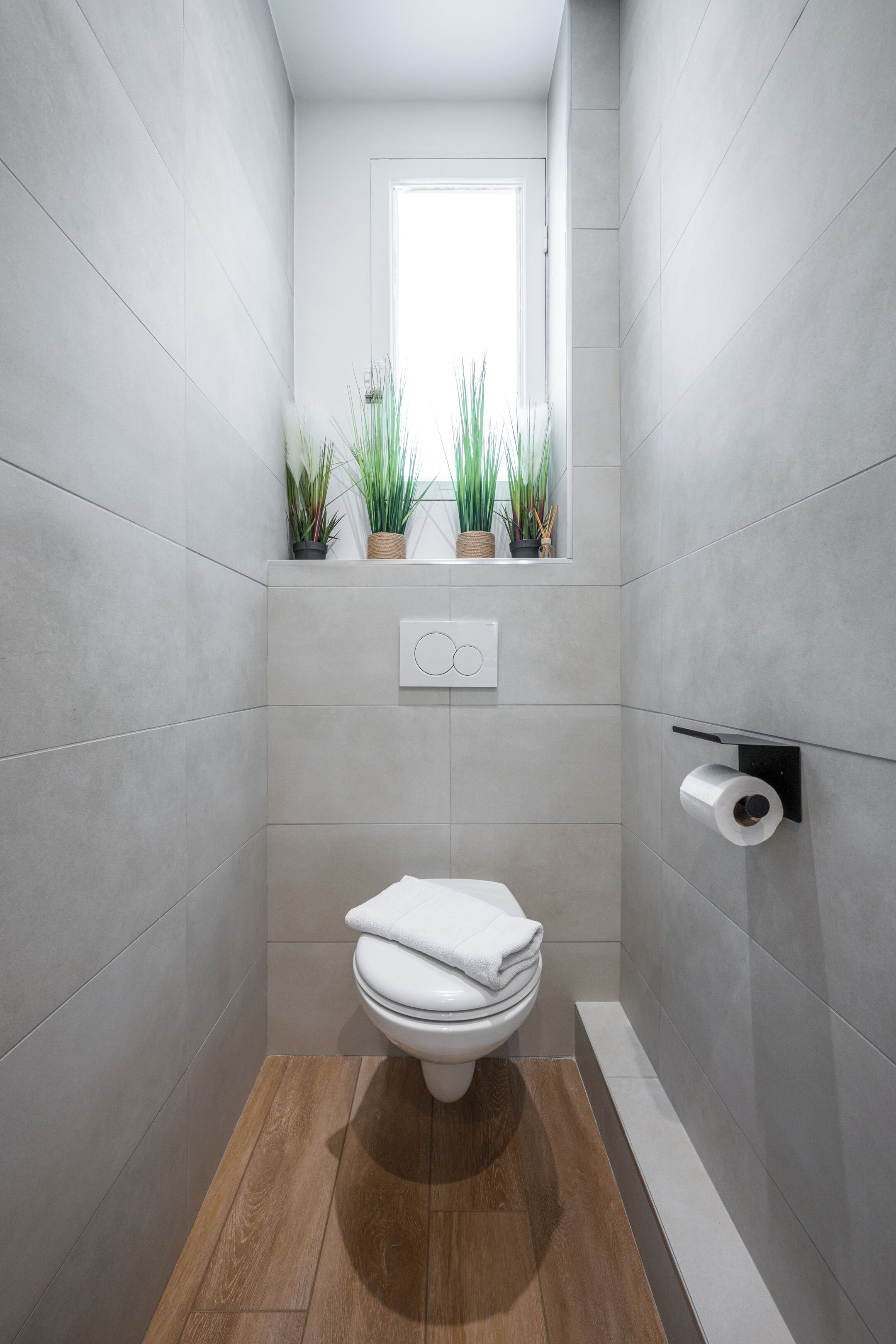 Un baño moderno con azulejos grises, suelo de madera, un inodoro, una ventana con plantas y un dispensador de papel higiénico de pared.