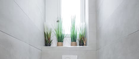Un baño moderno con azulejos grises, suelo de madera, un inodoro, una ventana con plantas y un dispensador de papel higiénico de pared.