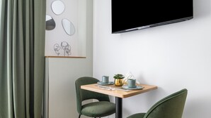 A modern room with a flat-screen TV mounted on the wall, two green chairs around a wooden table, and a shelf with decorative items. - L'App-Art (Nice)
