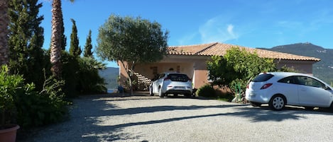 A house with two cars parked in front, surrounded by greenery and a clear sky.