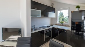A modern kitchen with black cabinets, a stainless steel refrigerator, a built-in oven, and a window providing natural light. - Two-bedroom apartmetn with sea view (Novalja)