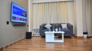 A modern living room with a flat-screen TV mounted on the wall, a grey sofa with cushions, a white coffee table with decorative items, and a wooden floor.