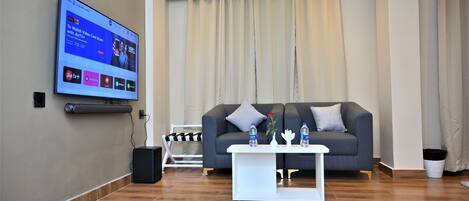 A modern living room with a flat-screen TV mounted on the wall, a grey sofa with cushions, a white coffee table with decorative items, and a wooden floor.