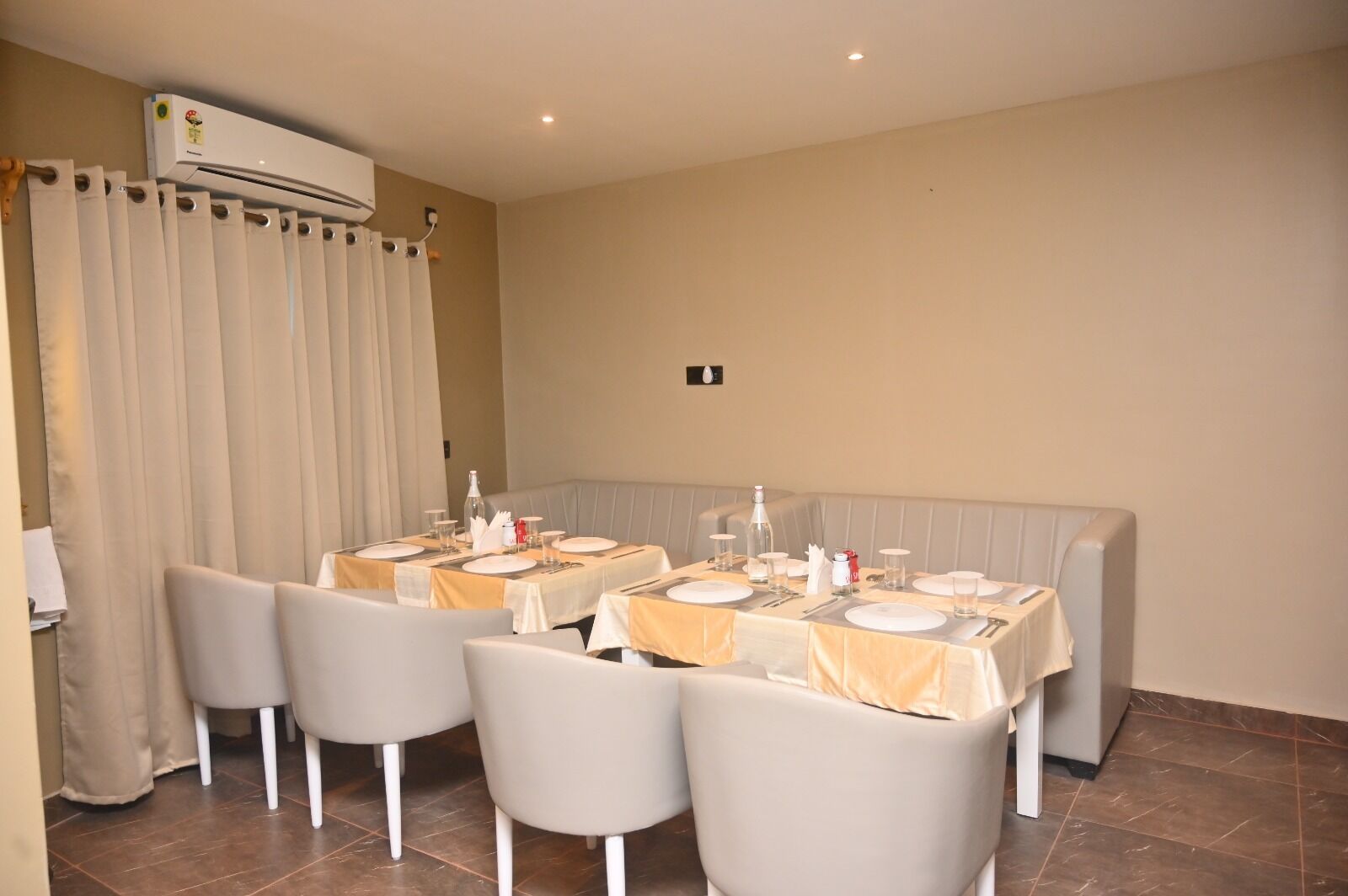 Un comedor con mesas dispuestas para cuatro, cada una con mantel blanco y cubiertos. Se observa un sofá color beige al fondo y un equipo de aire acondicionado en la pared.