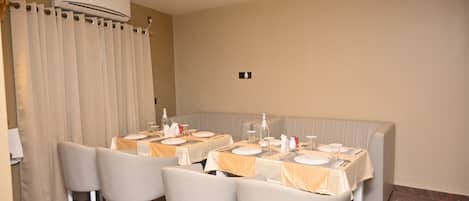 A dining area with tables set for four, each with white tablecloths and silverware. There is a beige sofa in the background and an air conditioning unit on the wall.