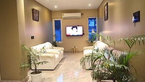 A living room with two white sofas, a flat-screen TV mounted on the wall, and several potted plants.