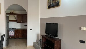 A living room with a flat-screen TV on a wooden stand, a kitchen visible through an archway, and a painting on the wall. - Casa Mim in Cabras (Cabras)