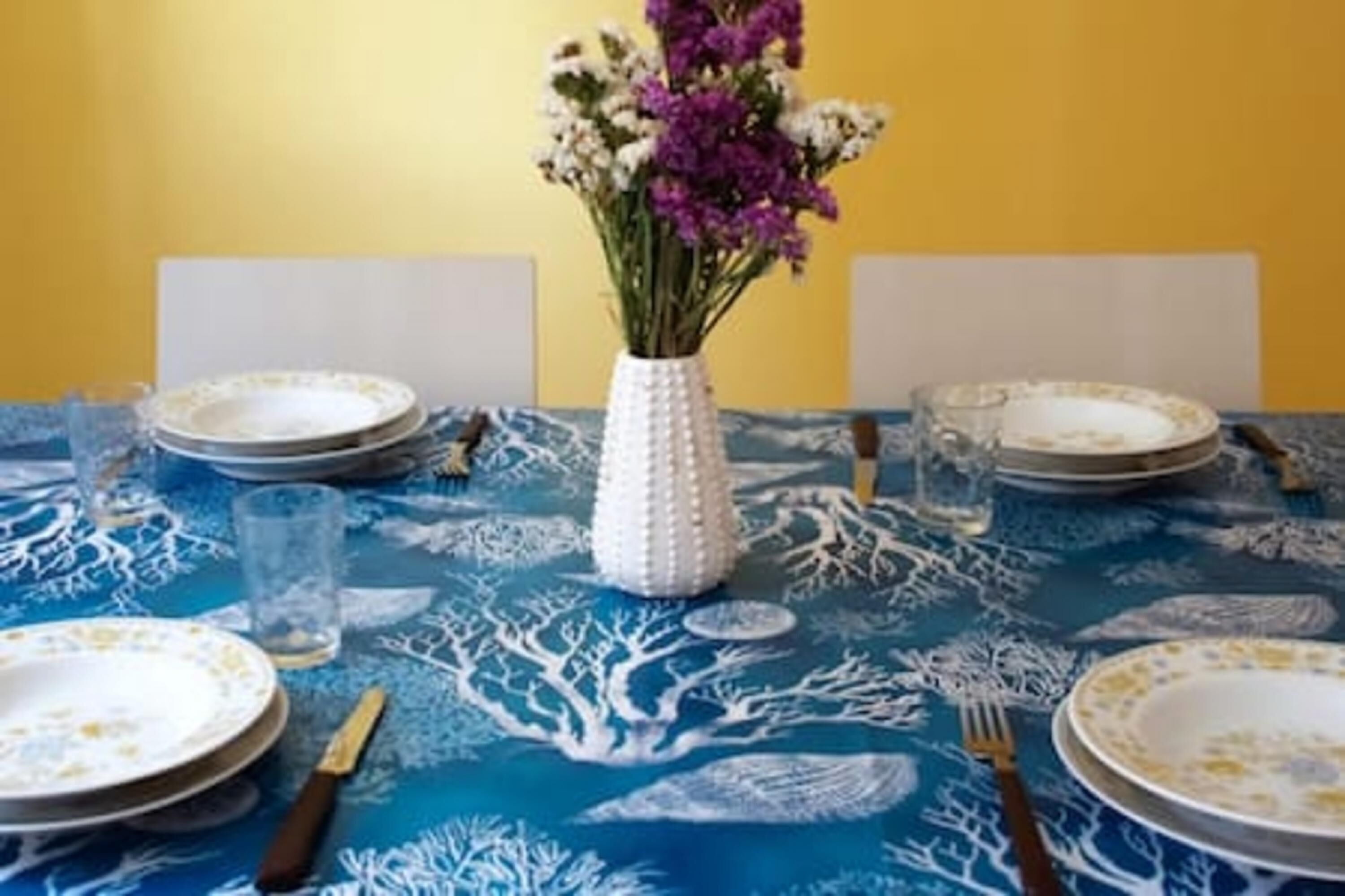 Mesa lista para comer con platos, cubiertos y un jarrón de flores frente a una pared amarilla.
