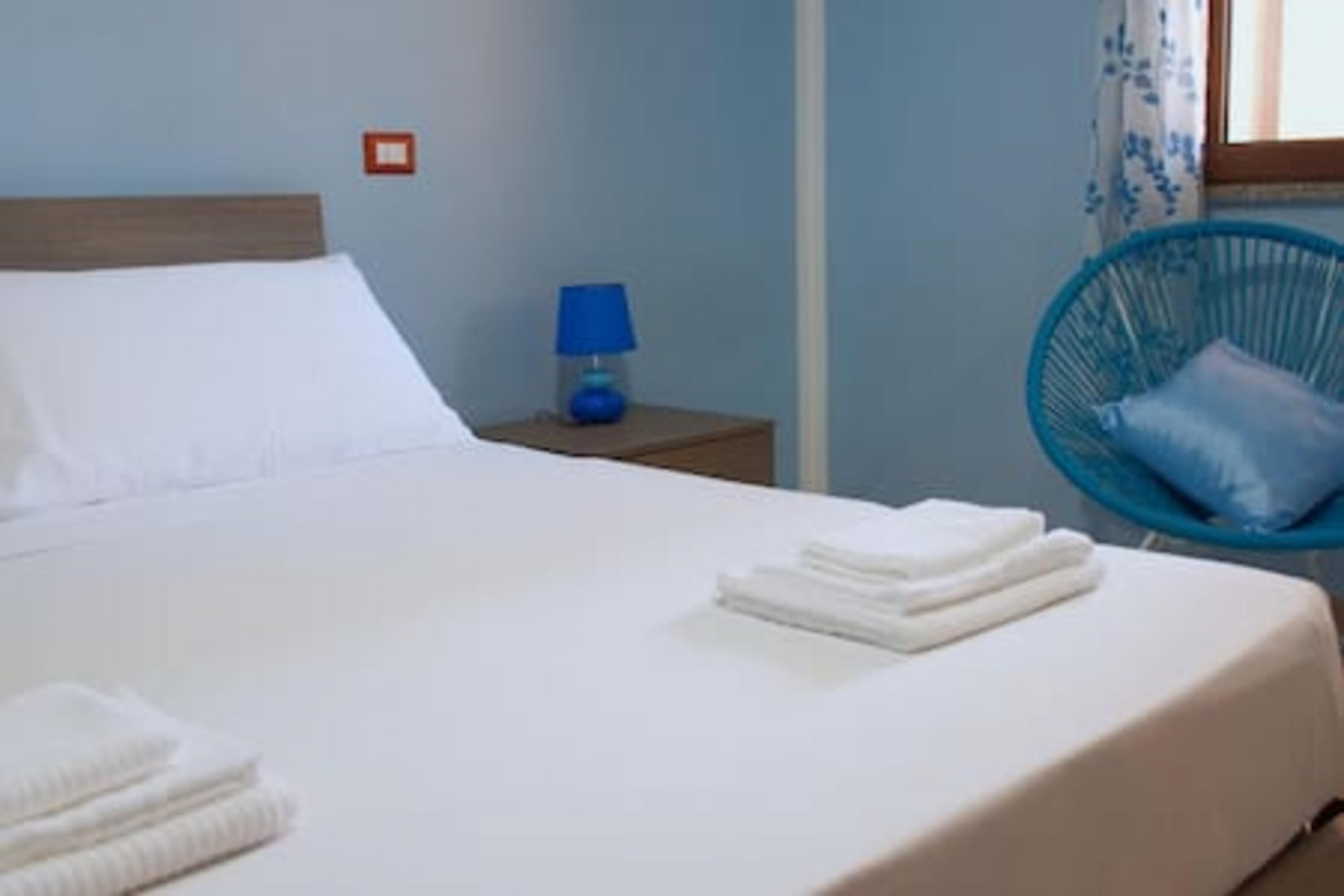 Ein ordentlich bezogenes Bett mit weißen Leinen und gefalteten Handtüchern, eine blaue Lampe auf einem Nachttisch und ein blauer Stuhl mit Kissen.