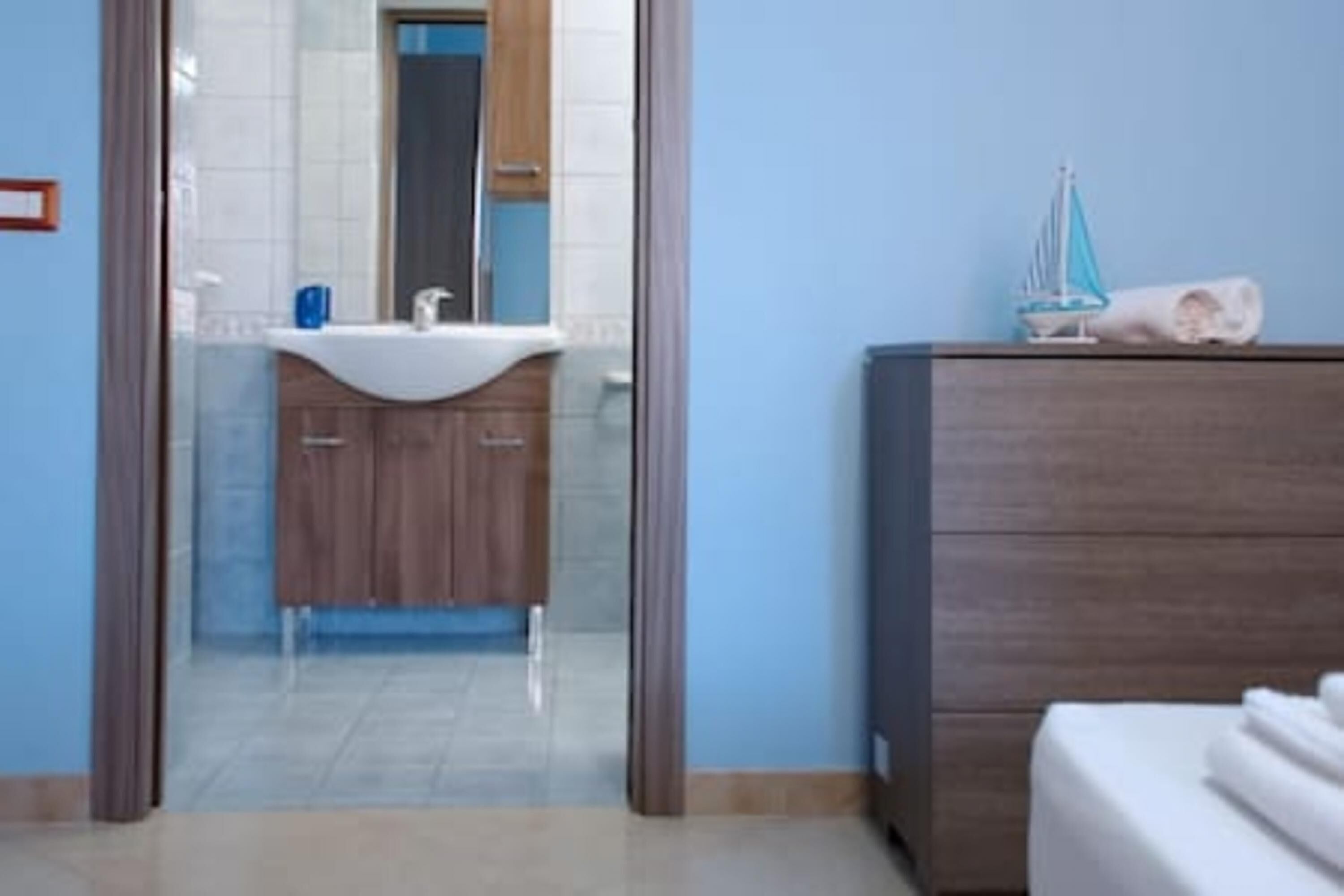 Un baño moderno con separador de vidrio, un lavamanos blanco, armarios de madera y un aparador de madera con una toalla y un pequeño barco modelo.
