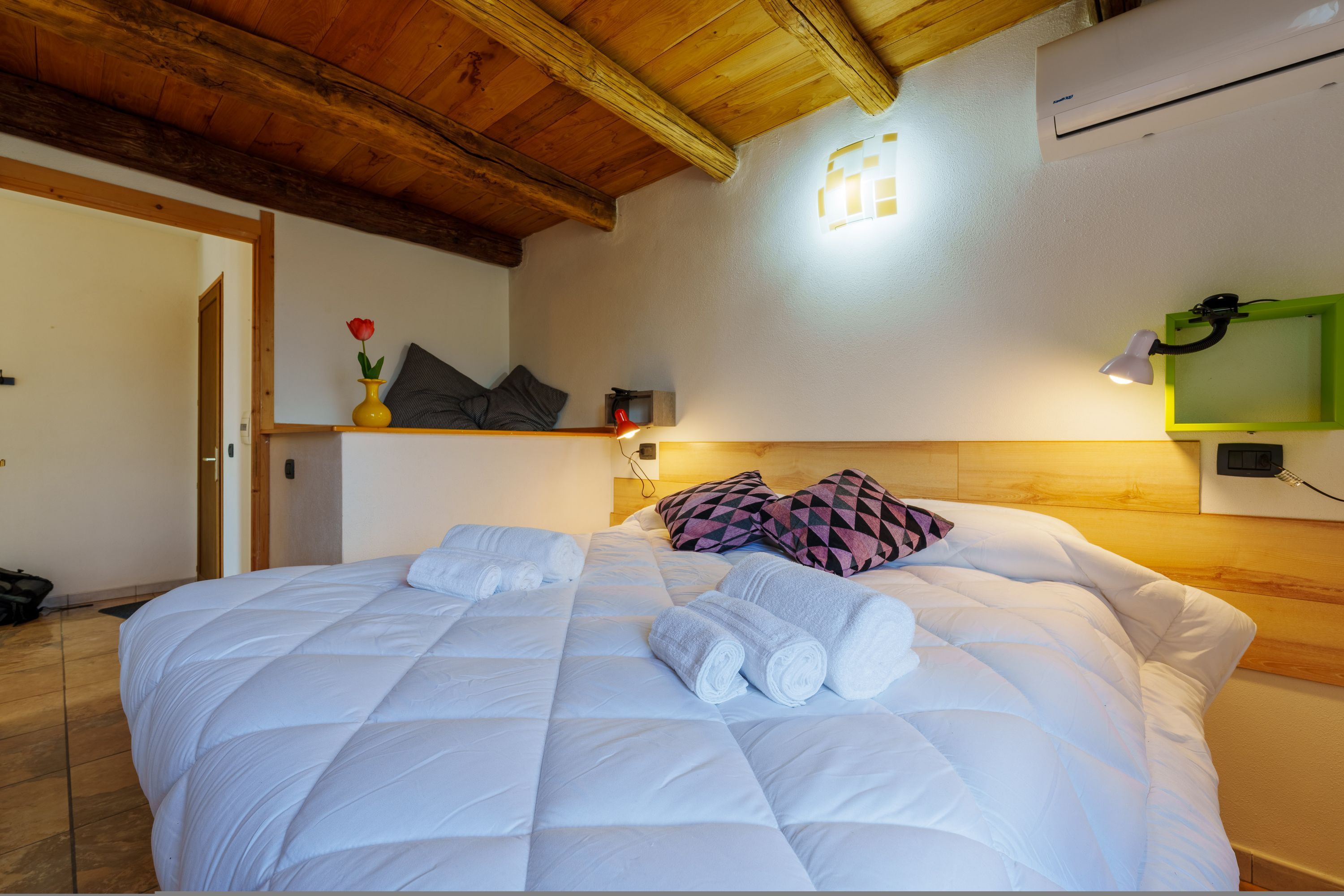 A neatly made bed with white bedding and rolled towels, a wooden ceiling, a wall-mounted air conditioner, and a green-framed mirror.