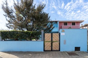 Une entrée fermée par un portail, avec un mur bleu, un arbre et un bâtiment à la façade rose.