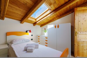 Une chambre avec un plafond en bois, un lit recouvert de draps blancs, une table de chevet, une commode avec des poignées orange et une armoire en bois.