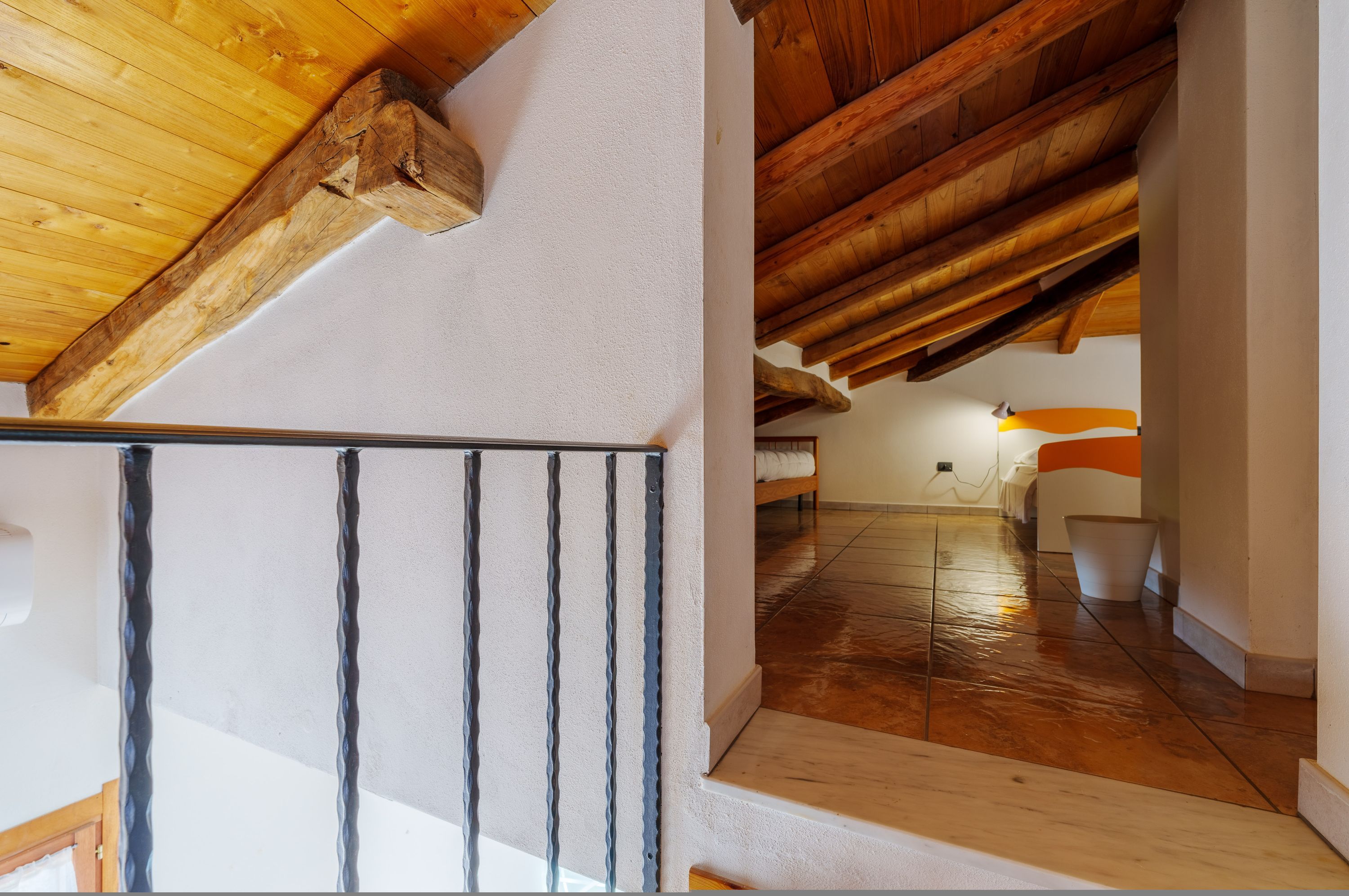 A wooden ceiling with exposed beams, a staircase with a metal railing, and a hallway leading to a room with a bed and a nightstand.