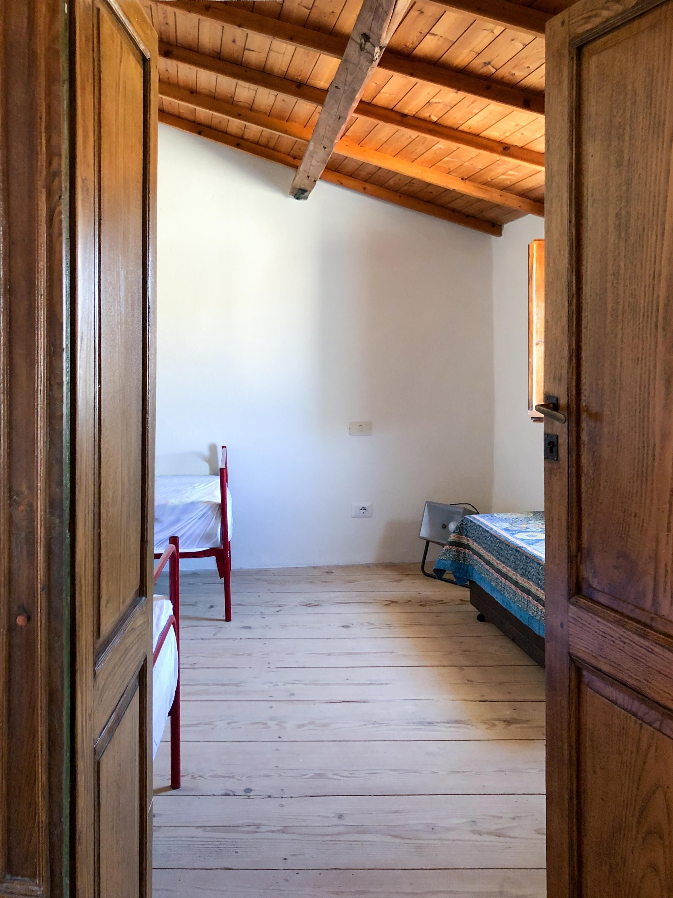 A room with wooden floors, a bed with a patterned bedspread, a chair, and a wooden door.